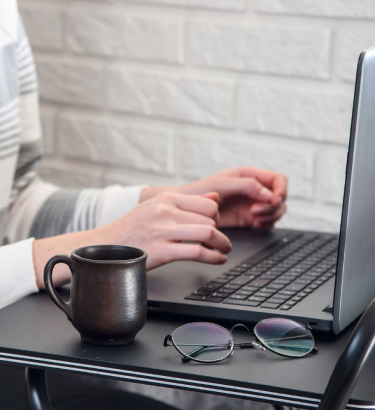 A person wearing glasses sits at a table, focused on their laptop, engaged in blog writing